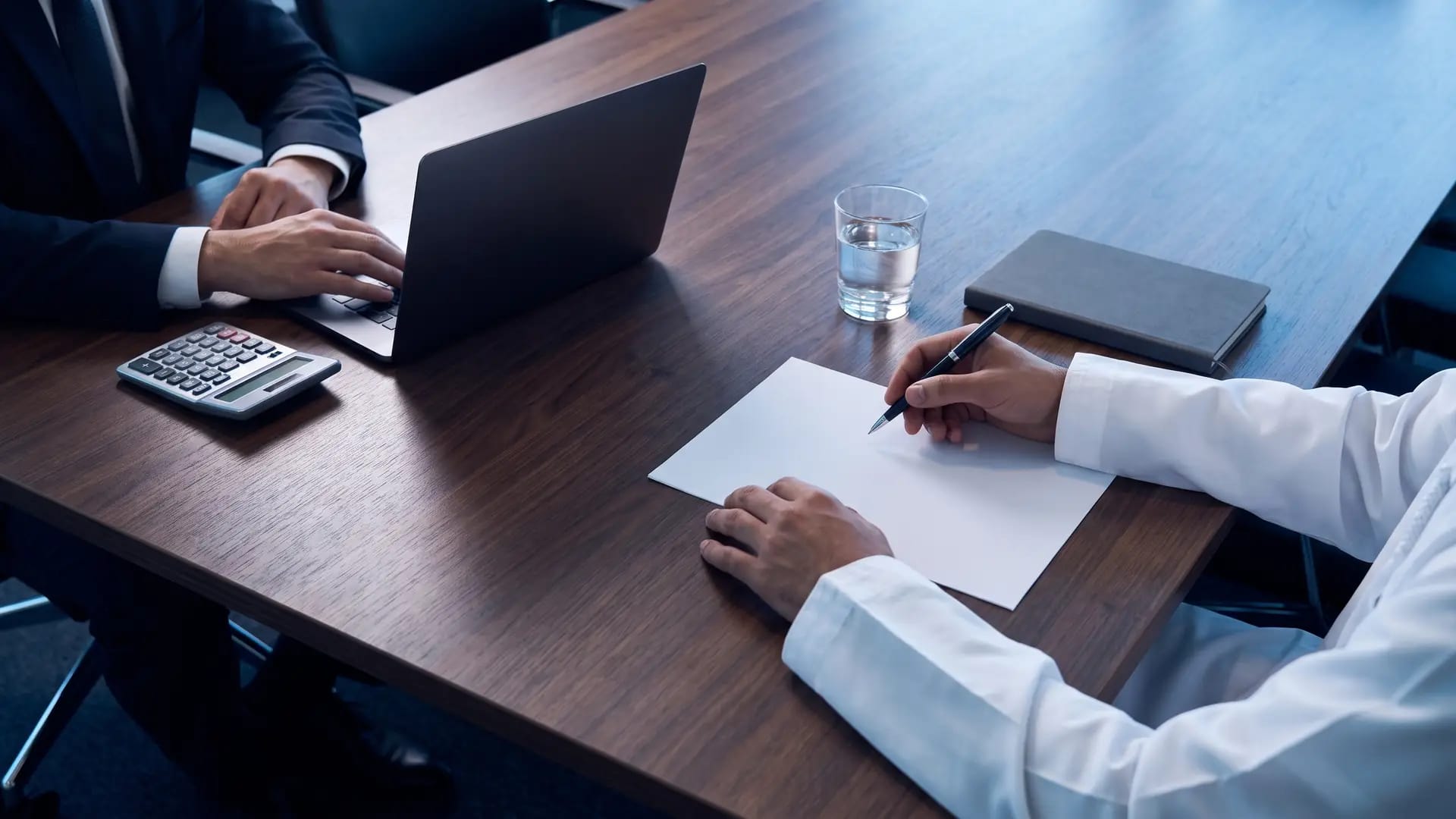 Two people at a dark wood meeting table working through a payroll calculation with a calculator, laptop and blank printed sheet