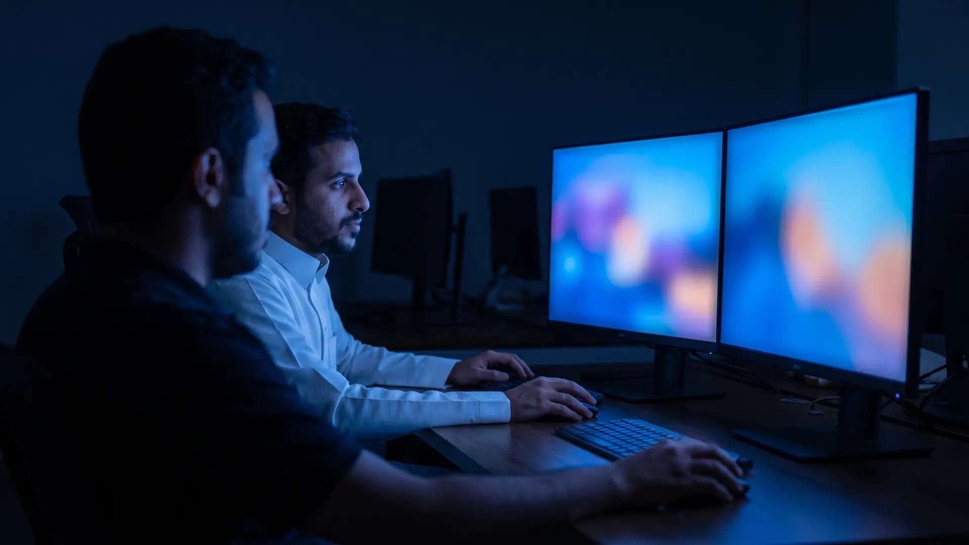 Two developers working side by side at a dual-monitor workstation in a darkened office, the screens glowing blue on their faces