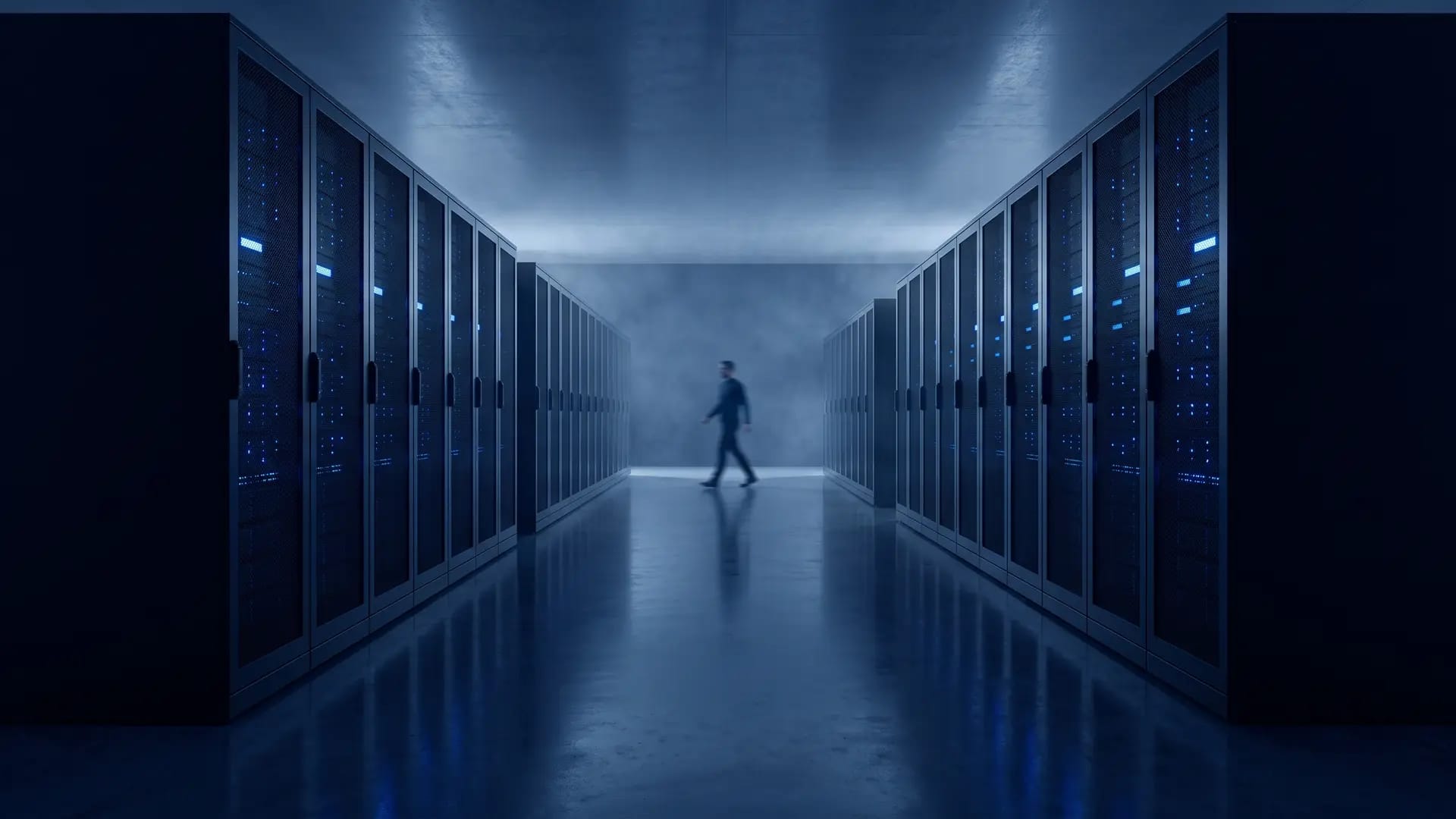 A wide aisle inside a data centre, rows of dark server racks lit by blue status lights with a technician walking away from camera
