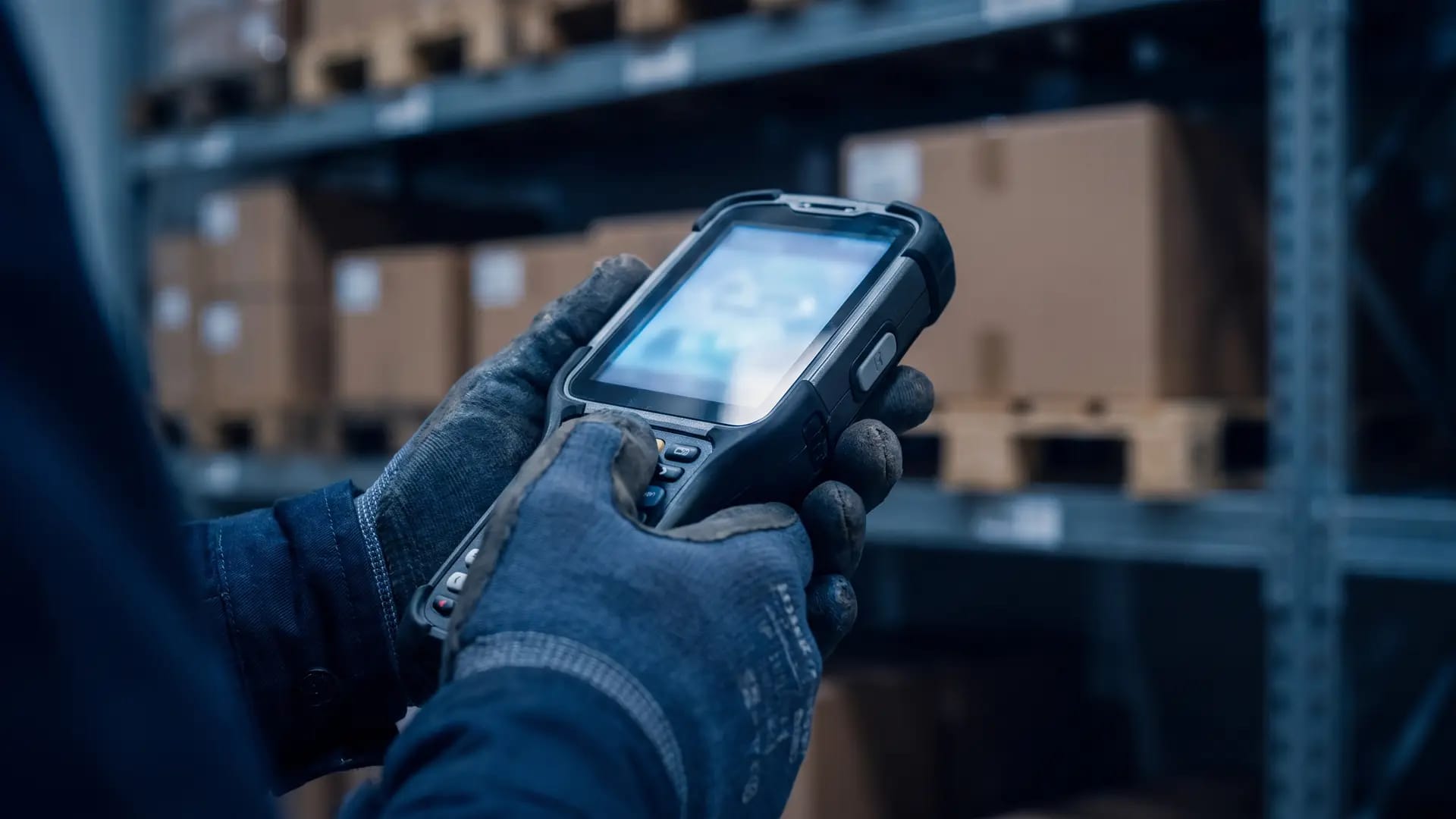 A gloved warehouse hand gripping a rugged RF barcode terminal with pallet racking blurred in the background