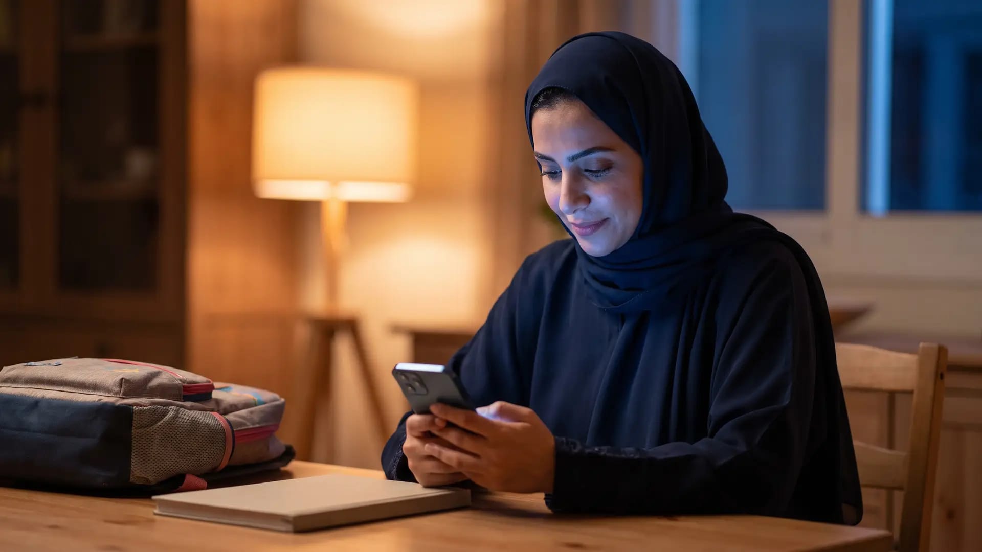A mother at a home dining table in the evening checking a smartphone, with a child's school backpack beside her