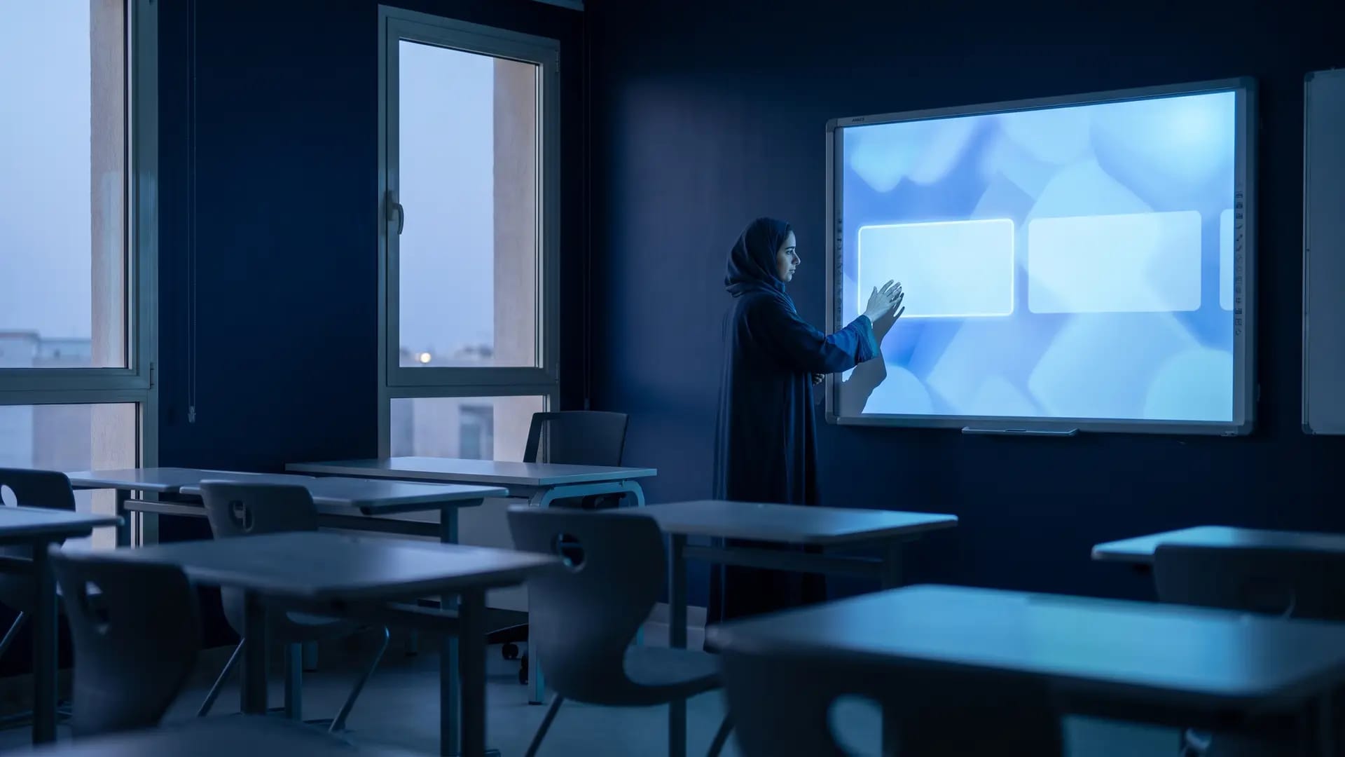 A teacher in a dark abaya standing beside a large illuminated interactive display in an empty modern classroom at dusk
