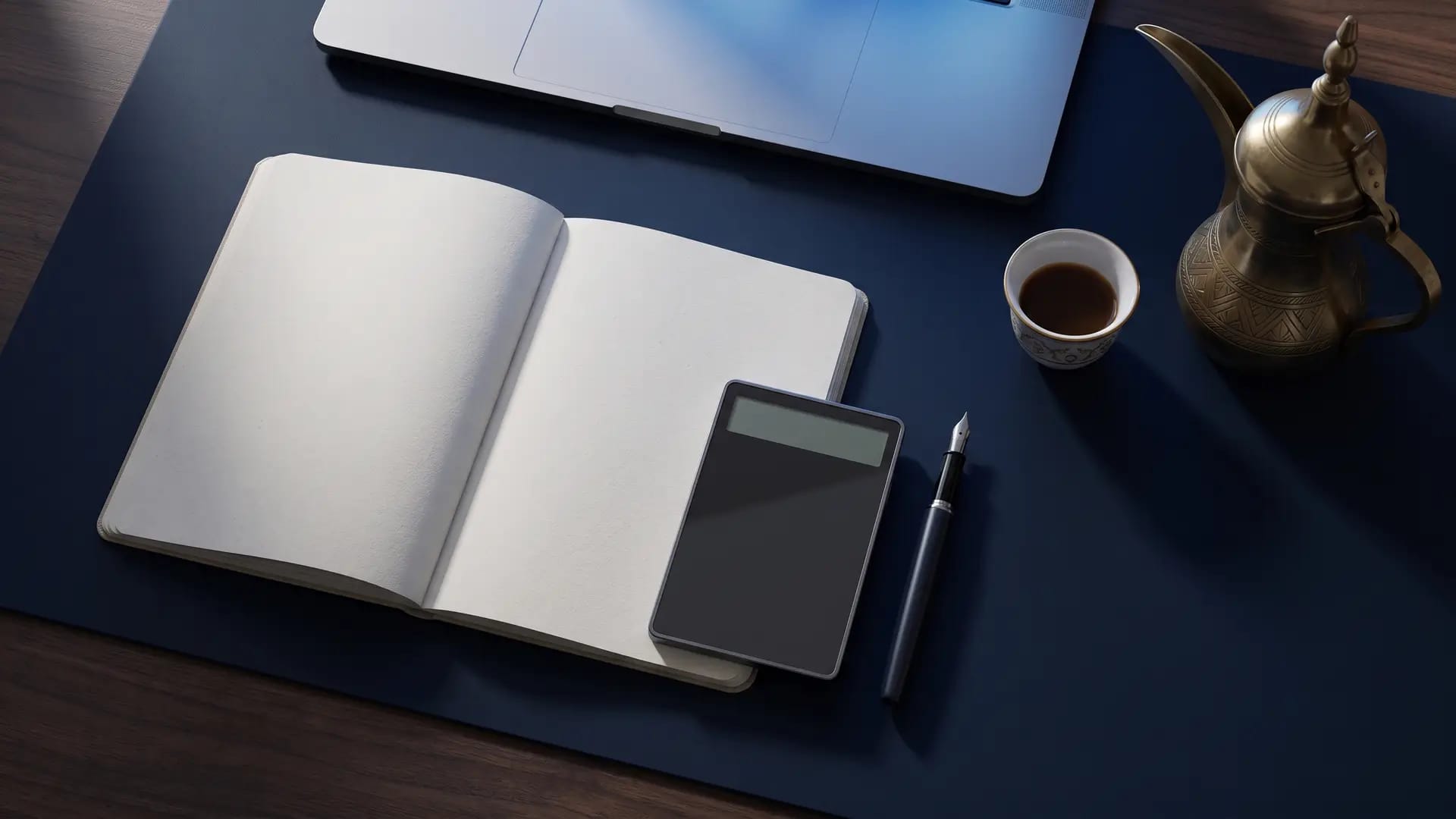 Overhead view of a dark wooden desk with a blank notebook, a calculator, a fountain pen and Arabic coffee beside a laptop
