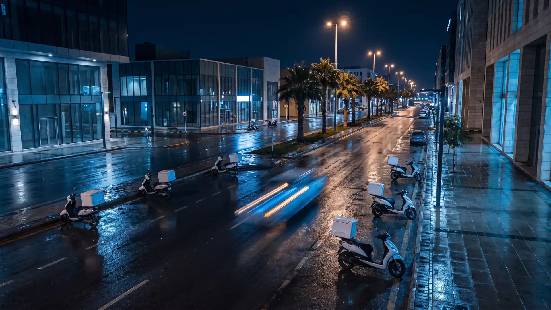 A wet modern Saudi city street at night with several parked delivery scooters carrying plain insulated boxes and one moving vehicle blurred by motion