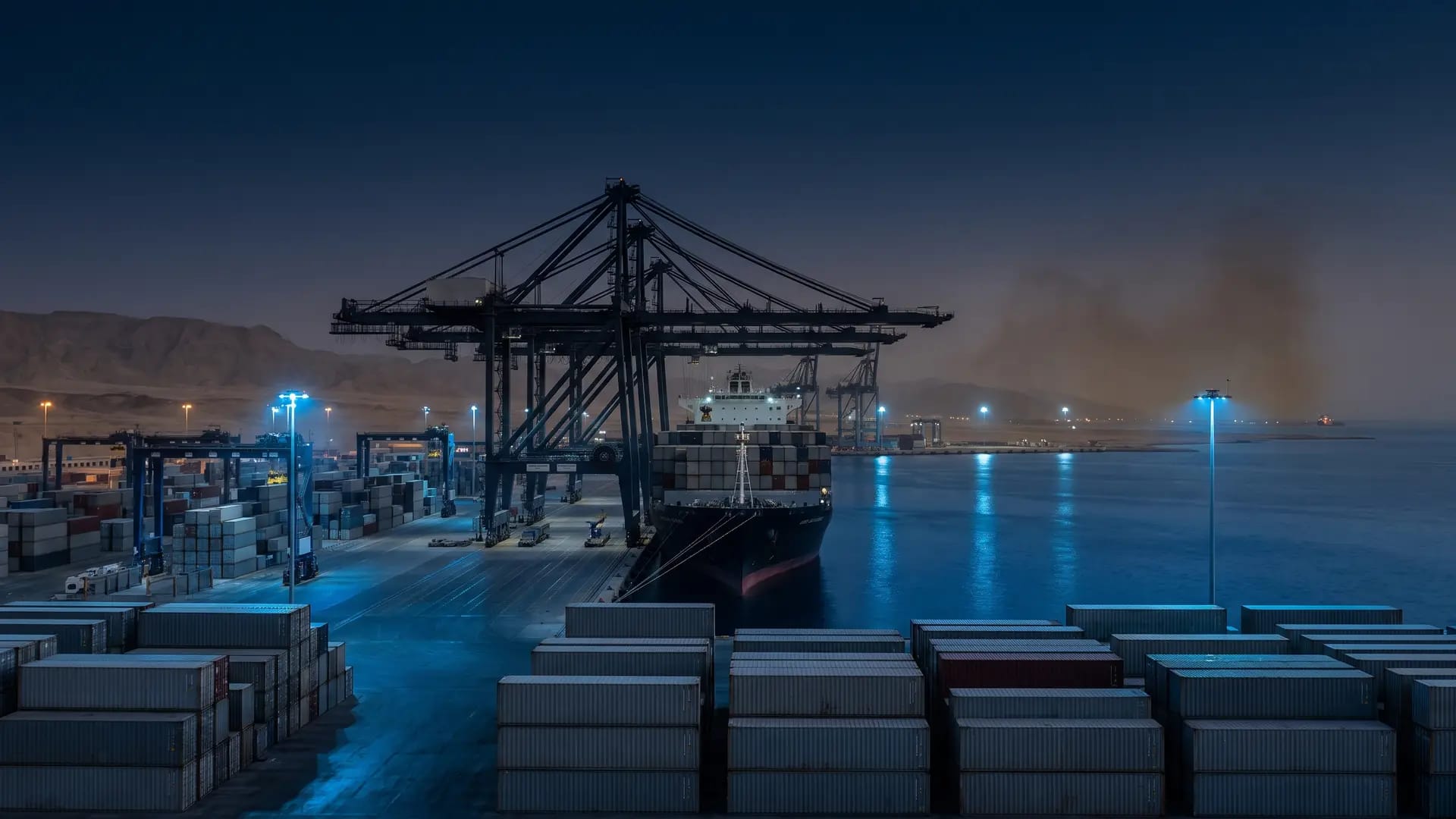 Ship-to-shore gantry cranes and stacked shipping containers at a Red Sea container terminal at blue hour, a container vessel moored alongside the quay