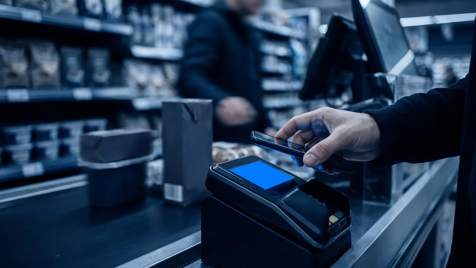 A shopper holding a smartphone above a contactless payment reader at a supermarket checkout, queue blurred behind