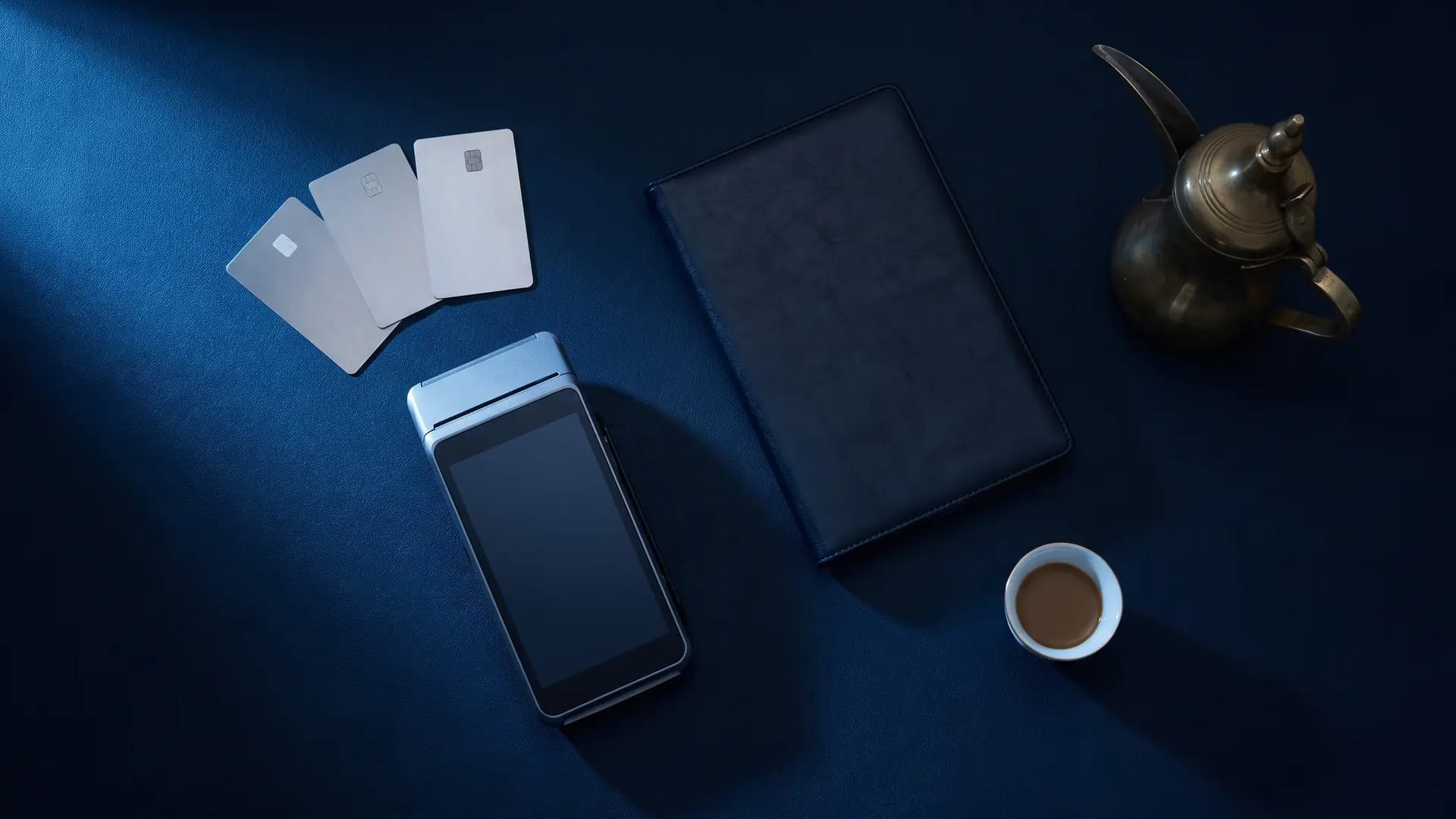 Overhead flat-lay of three unbranded payment cards, a blank-screened card terminal, a notebook and Arabic coffee on a dark navy desk