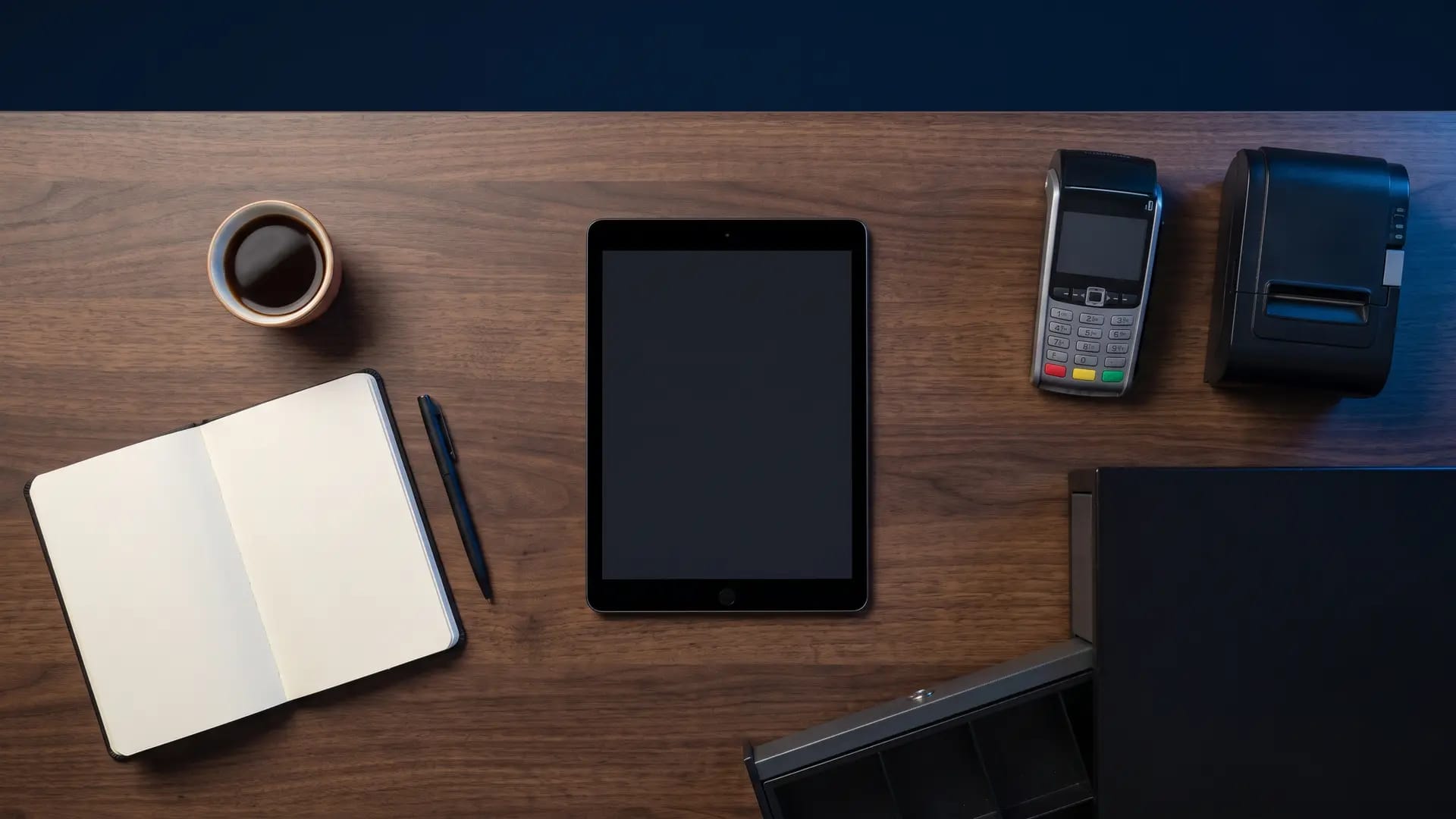 Overhead flat lay on a dark walnut desk with a tablet, a card payment terminal, a receipt printer, a cash drawer, Arabic coffee and a blank notebook