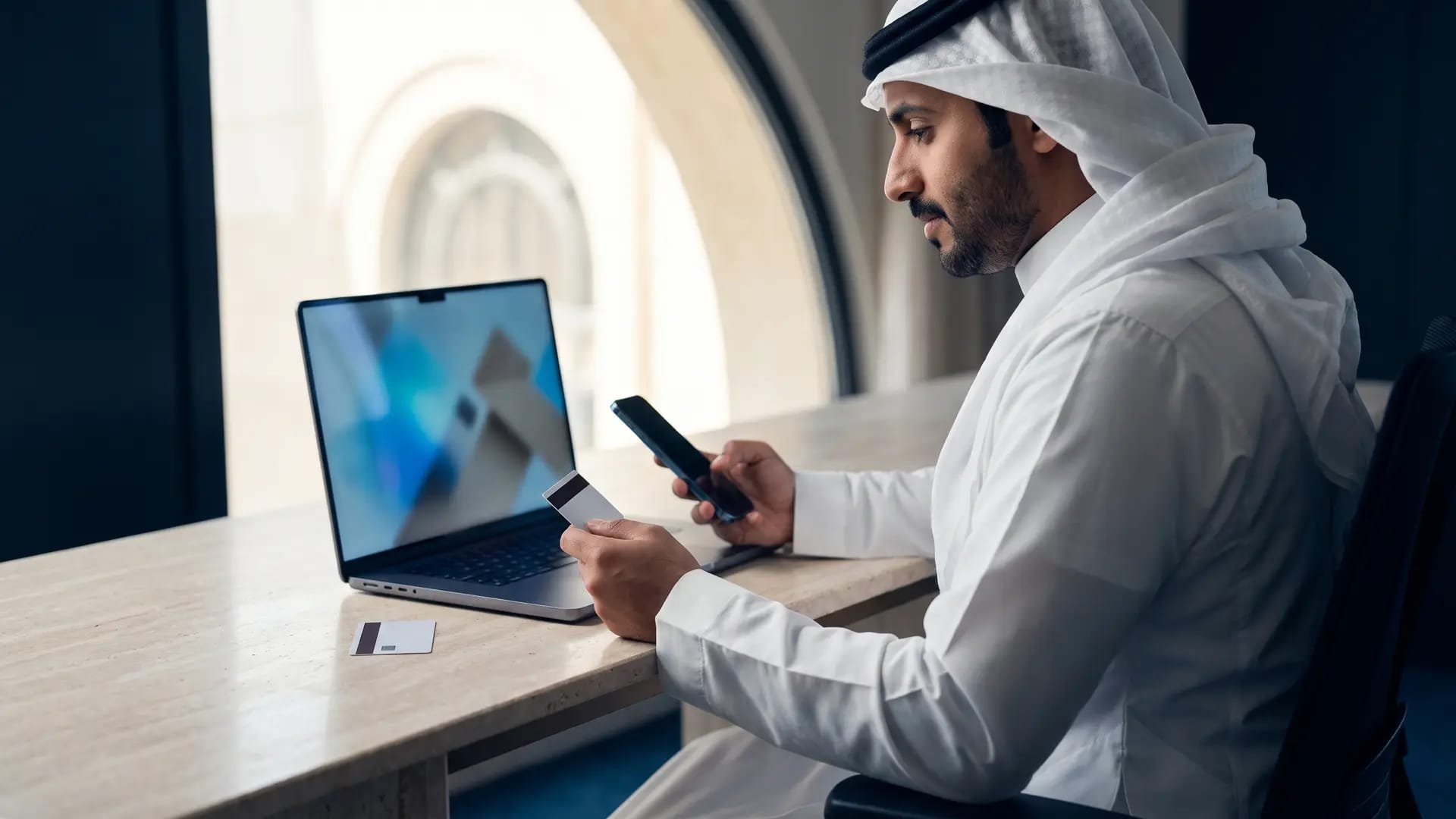 A Saudi business owner completing an online payment on a laptop and phone in a Riyadh workspace