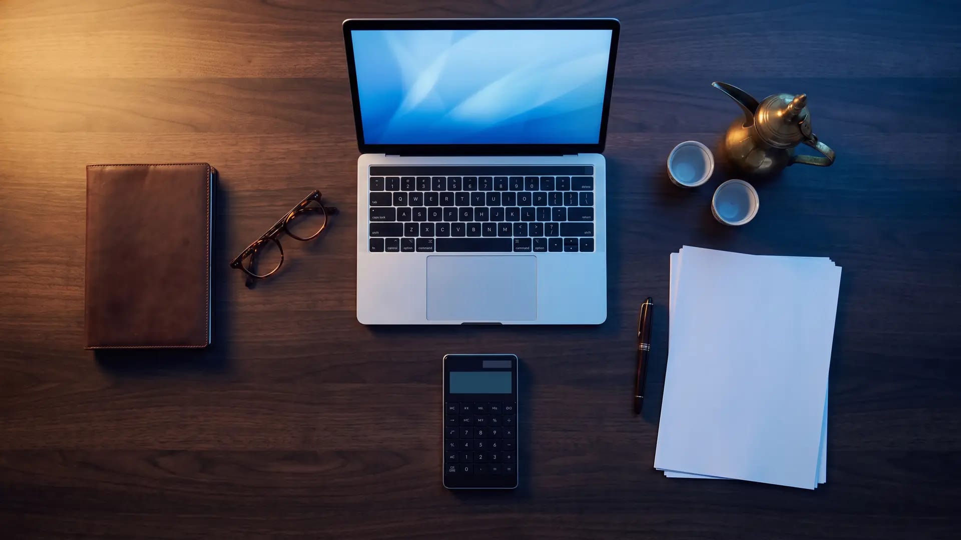Overhead view of a dark wood desk with a laptop, calculator, notebook and Arabic coffee pot during a cost review