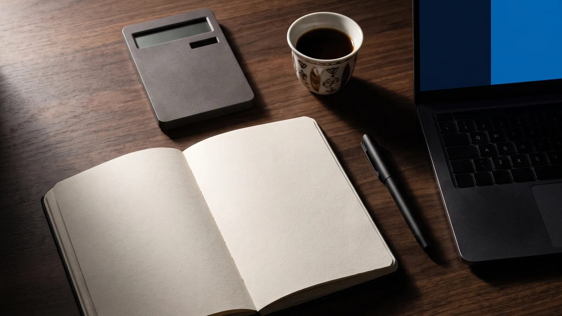 Overhead view of a dark wood desk with a notebook, calculator, Arabic coffee and a laptop showing a cost comparison