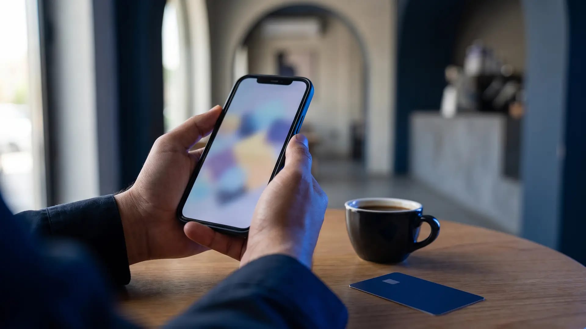 A Gulf shopper's hands holding a smartphone at a cafe table with a plain payment card and a cup of Arabic coffee beside it