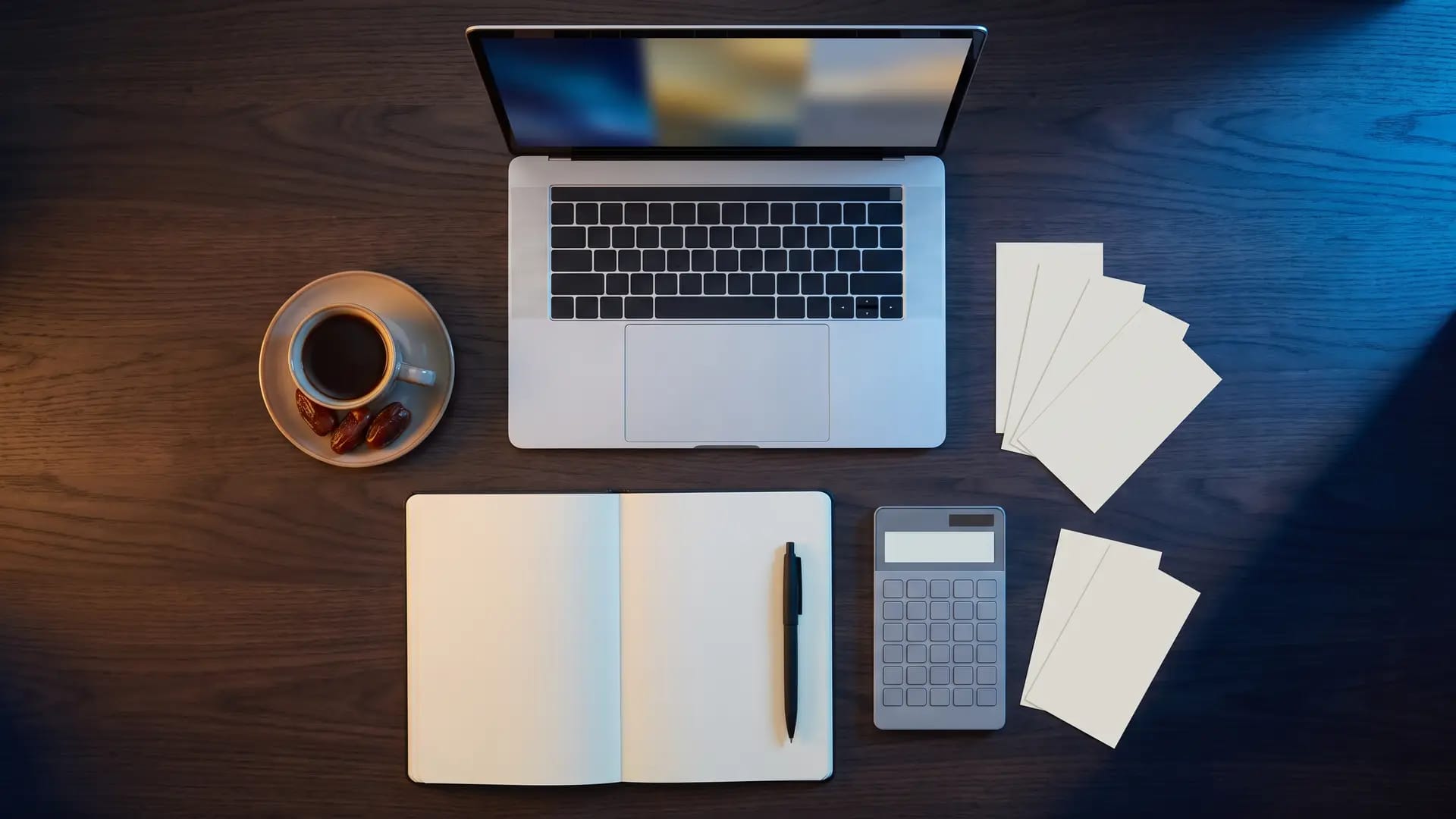 Overhead view of a dark wood desk with a blank notebook, a calculator, blank receipt slips and a cup of Arabic coffee beside a laptop