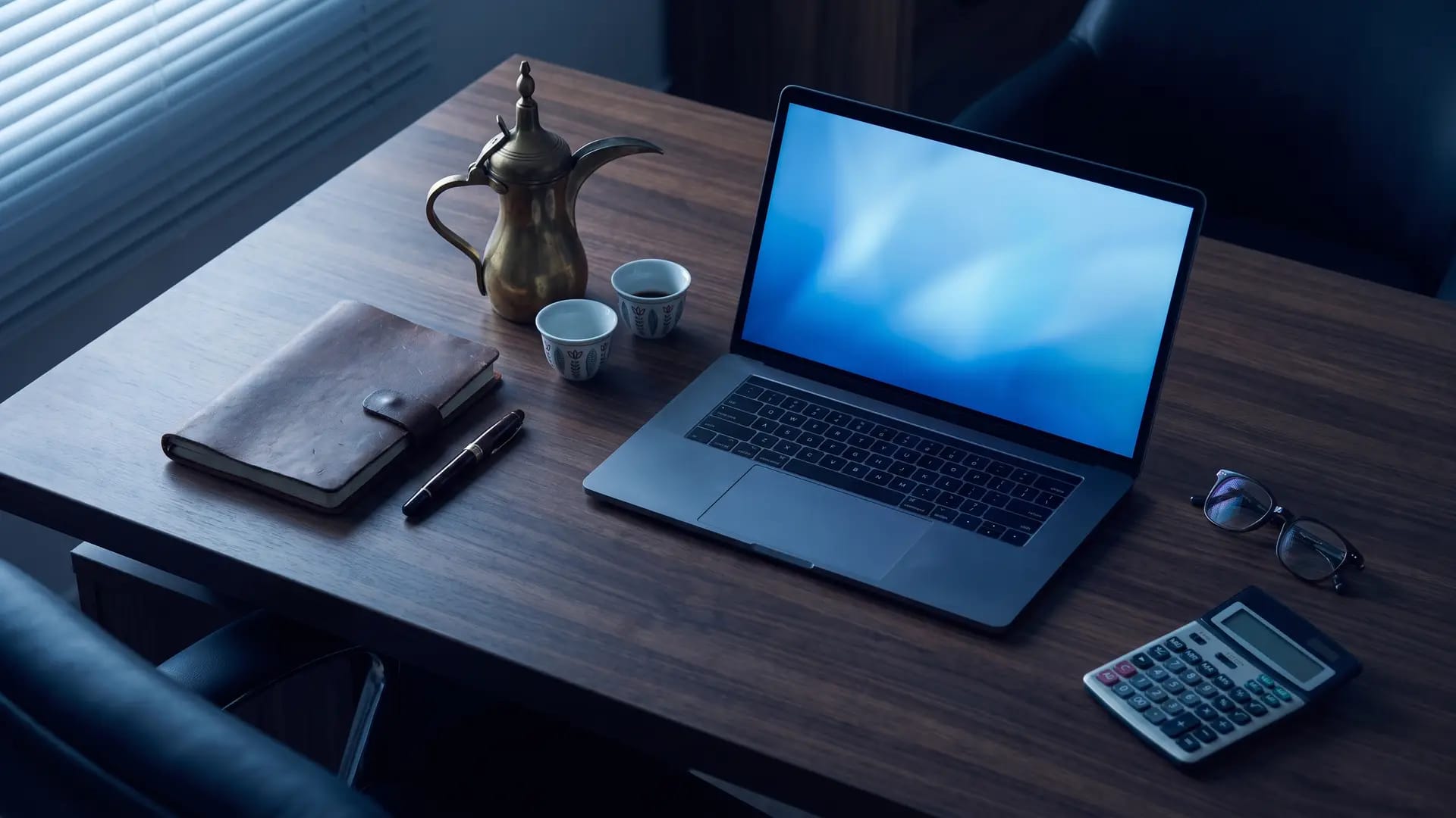Dark wood desk with an open laptop, a calculator, a leather notebook and an Arabic dallah coffee pot during an evening cost review