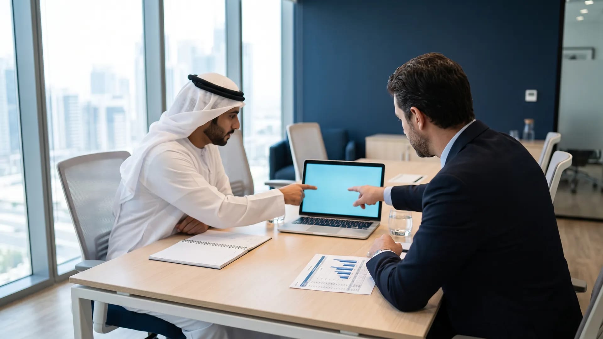 Two business colleagues, one in a white thobe and one in a suit, reviewing software options together on a laptop in a modern Gulf meeting room