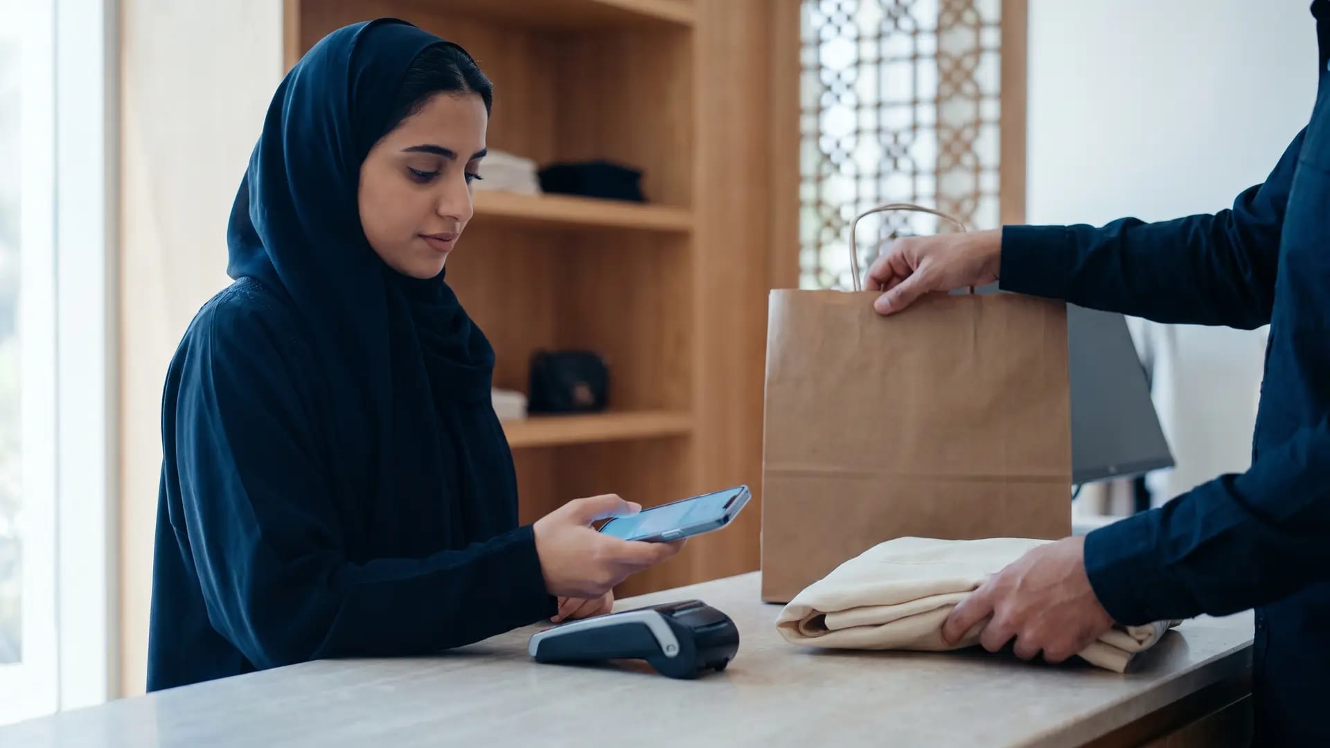 A shopper at a minimalist Saudi boutique counter holding a smartphone above a card terminal while an assistant folds a garment into a paper bag