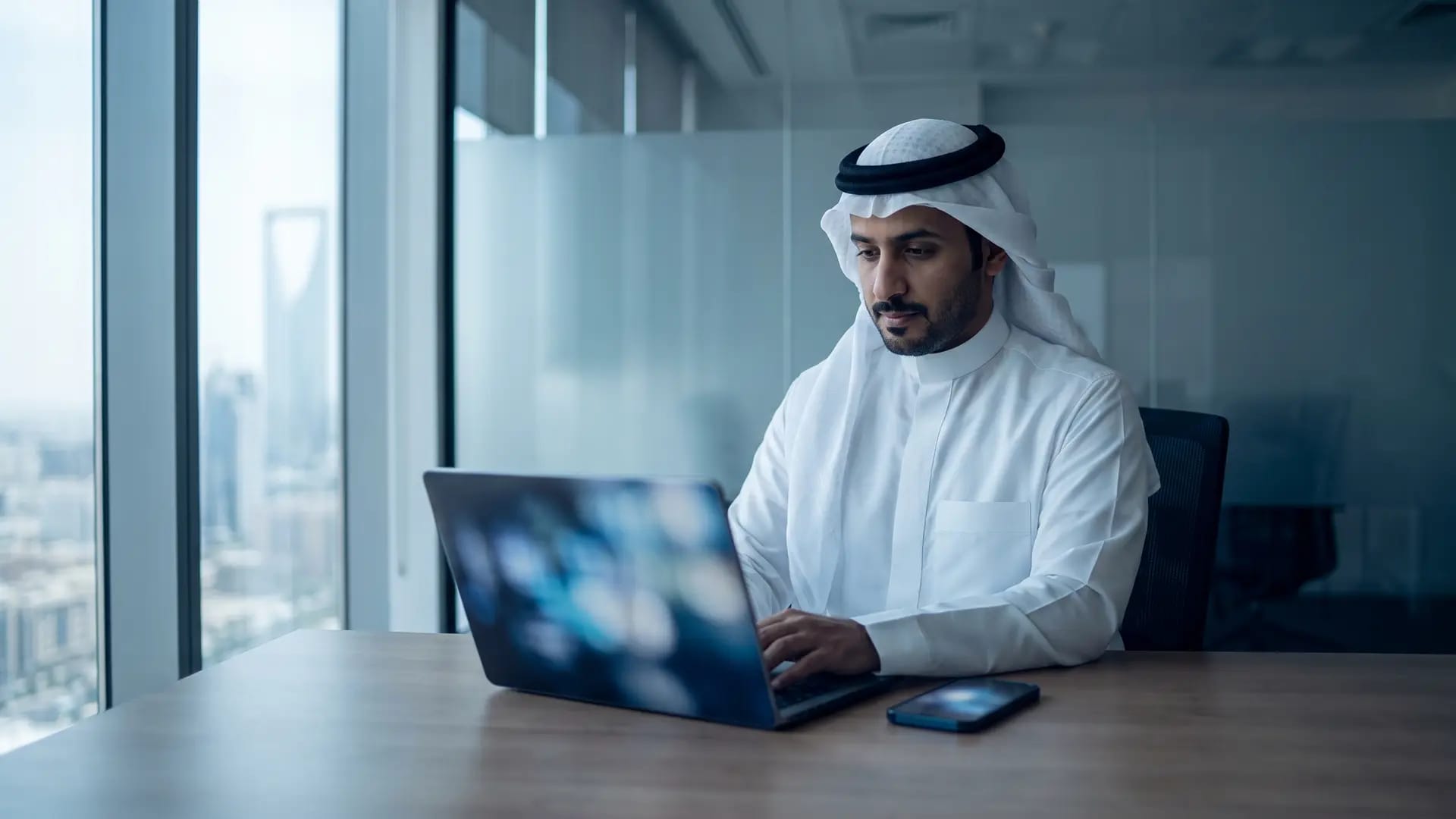 A Saudi business owner in a white thobe reviewing accounts on a laptop in a Riyadh office with the city skyline behind him