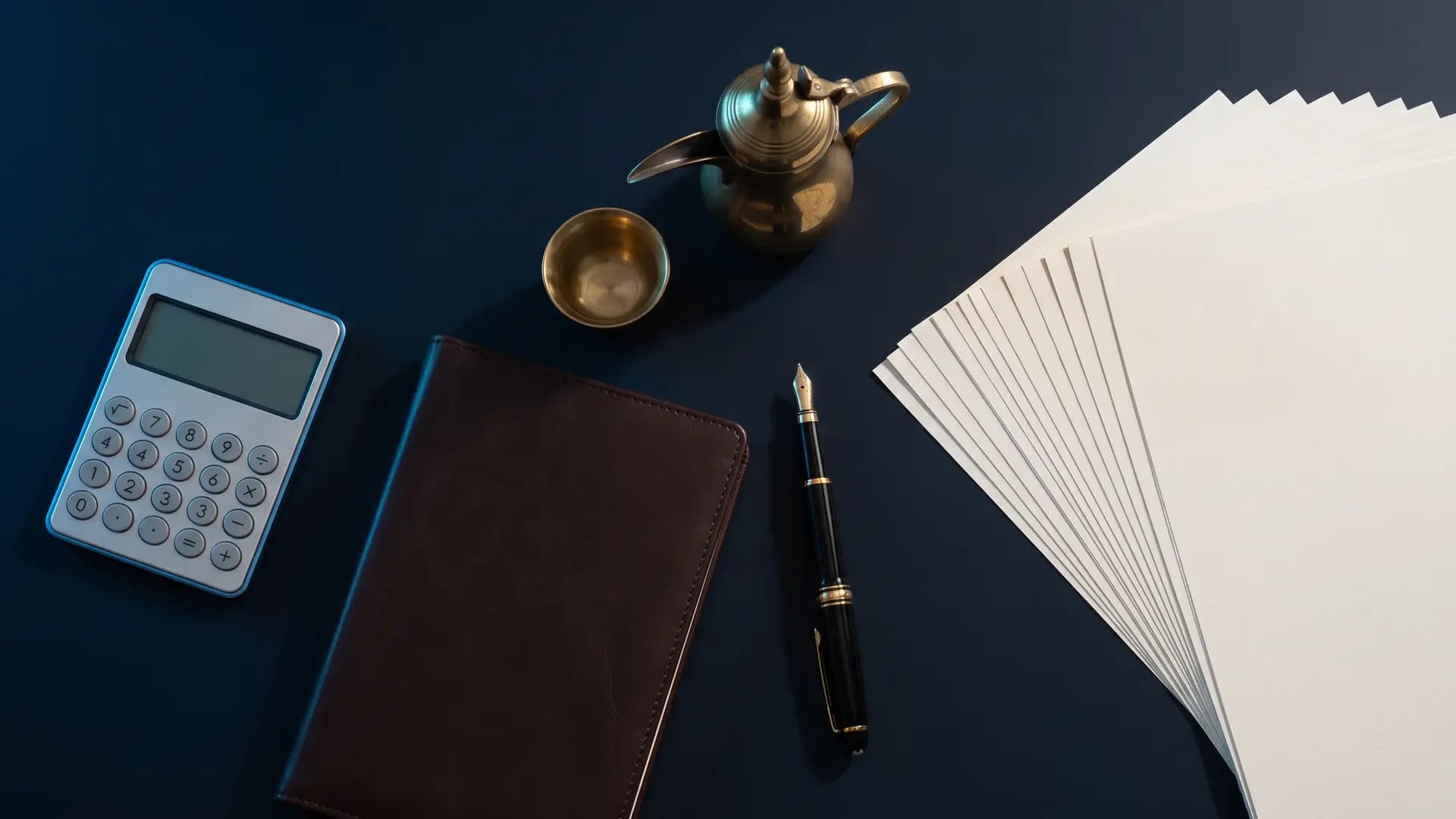Overhead flat lay on a navy desk with a calculator, leather notebook, fountain pen, brass Arabic dallah coffee pot and a fan of blank paper