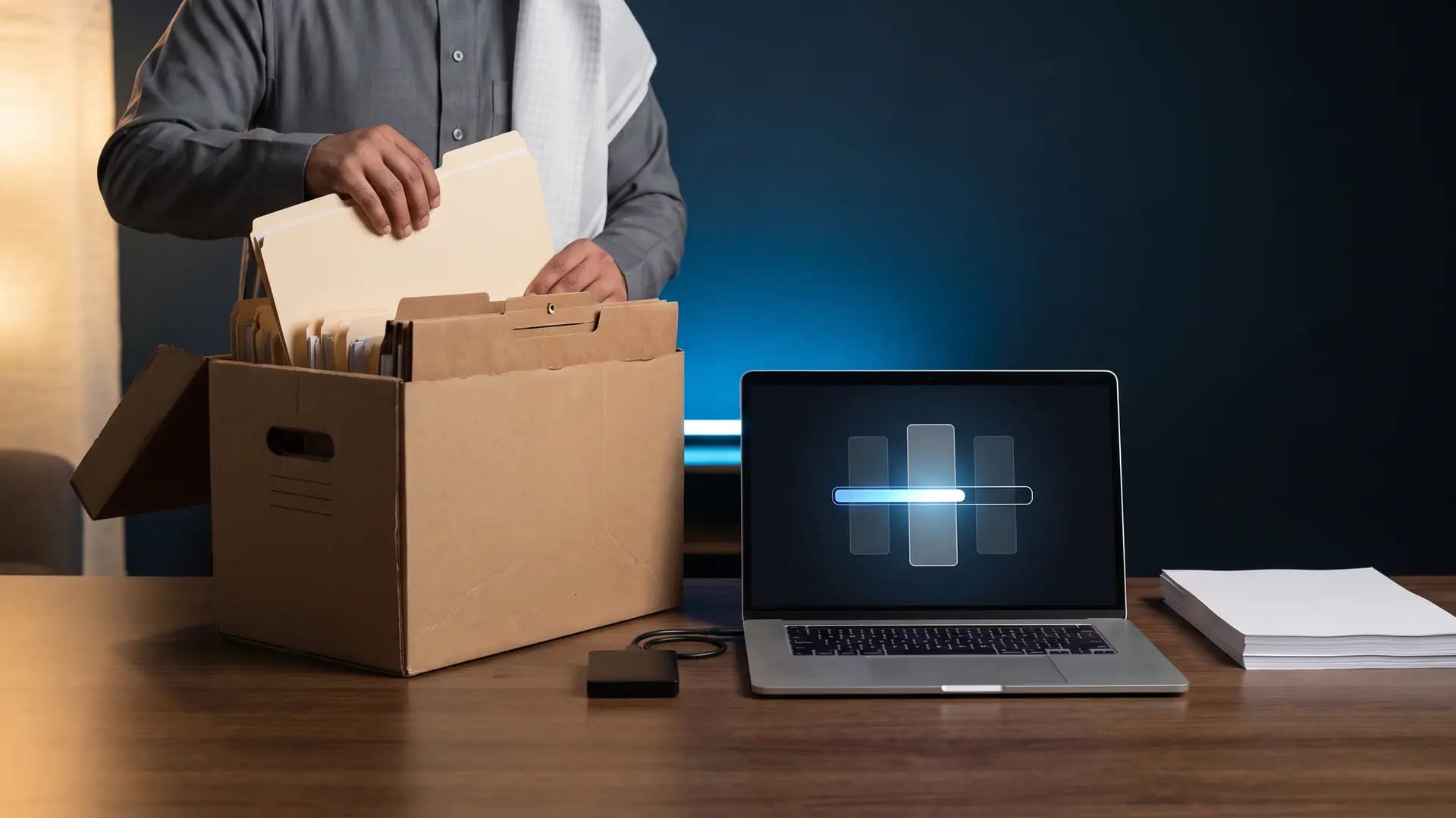 An accountant lifting a manila folder from an archive box beside a laptop showing a data transfer in progress