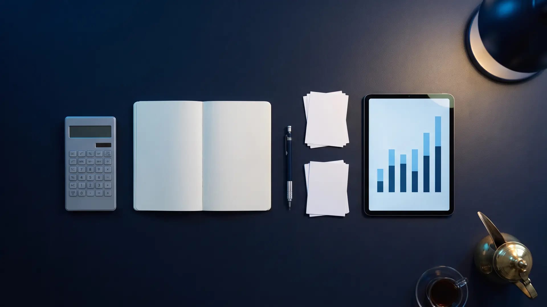 Overhead flat-lay of a navy desk with a calculator, blank notebook, paper slips and a tablet showing a bar chart, lit by a single desk lamp